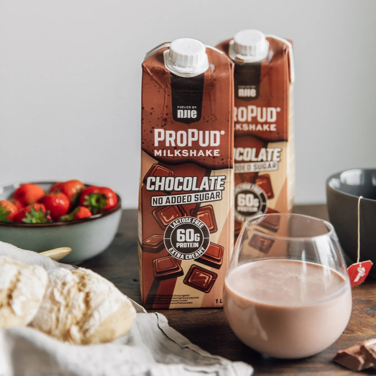 Two cartons of Propud Milkshake Chocolate No Added Sugar milkshake displayed on a rustic wooden table with a glass of the light brown shake, strawberries, and bread in the background.