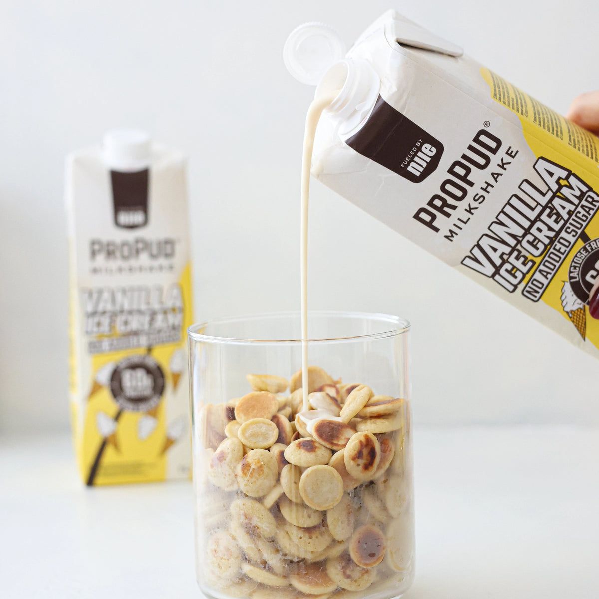 A hand holds a carton of Propud Milkshake Vanilla Ice Cream pouring the creamy beige liquid into a clear glass filled with cookies, with another Propud carton blurred in the background on a white surface.