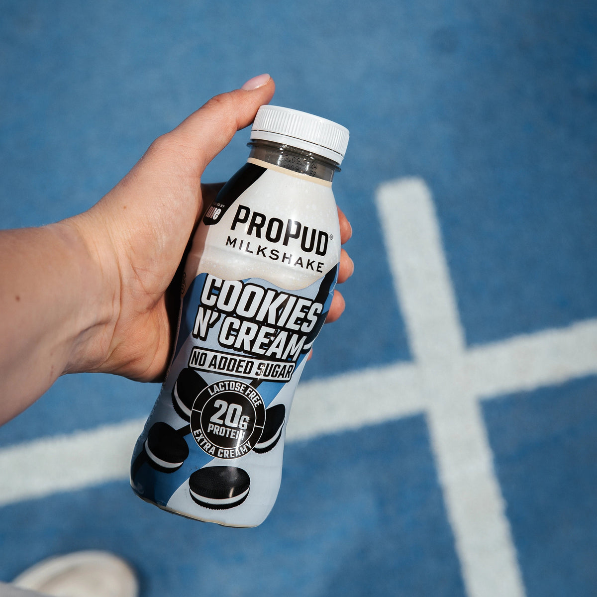 A hand holding a bottle of PROPUD MilkshAKE Cookies N Cream protein drink, set against a blue running track background with white lane markings and white sneakers visible at the bottom.