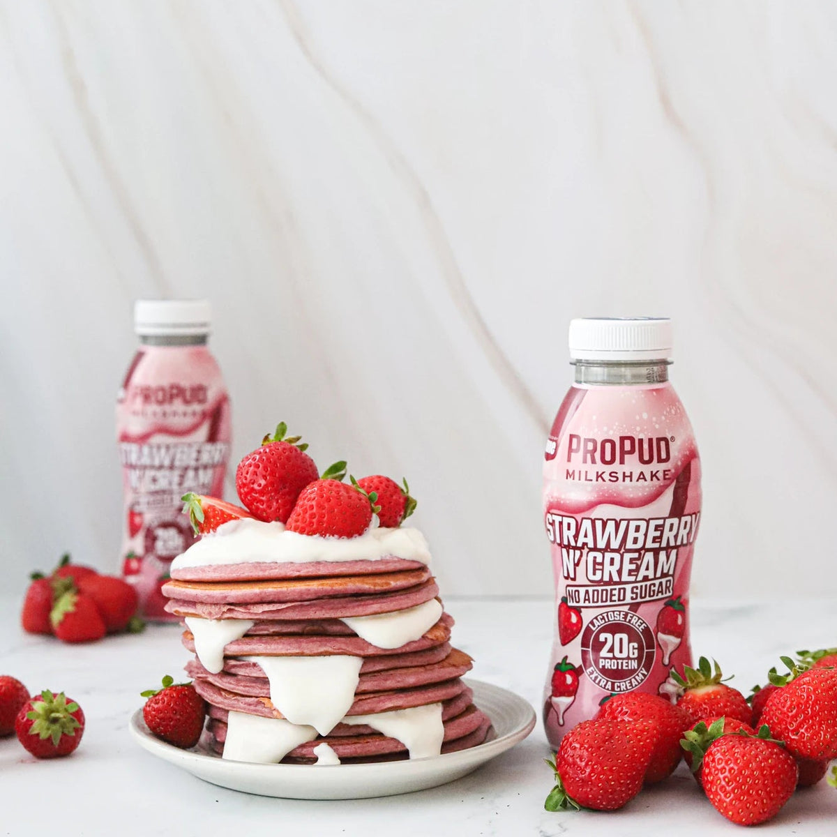 A styled product shot of Propud Milkshake Strawberry Cream flavored milkshakes. The foreground features a tall stack of strawberry pancakes with white whipped cream and fresh strawberries on a white plate, flanked by two bottles of the pink drink and scattered strawberries on a white marble surface.